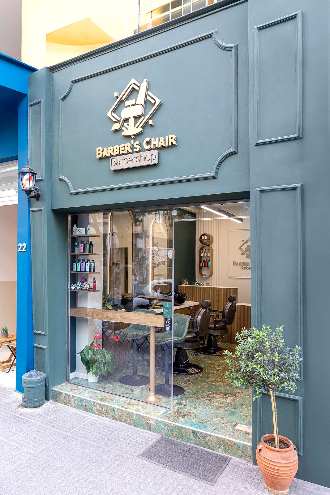 Η πρόσοψη του Barber's Chair στην Αλεξάνδρου Σβώλου 22: βαμμένη πρόσοψη σε βαθύ πετρόλ με χρυσό σήμα στο υπέρθυρο, τζαμαρία και μια ελιά σε γλάστρα στο πεζοδρόμιο